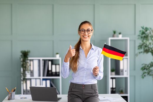 Frau mit Deutschlandfahne in der Hand vor Schreibtisch im Büro – Geförderte Deutschkurse: Mit einem Einzeltraining zu schnelle Erfolg!