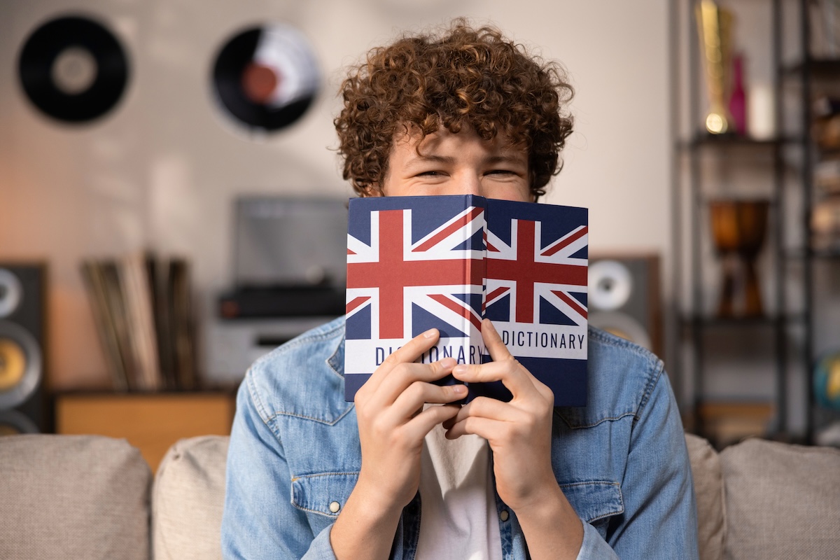 Teenager mit Locken hält sich einen Dictionary vors Gesicht – Gutes Business-Englisch ist eine Voraussetzung für viele Karrieren.